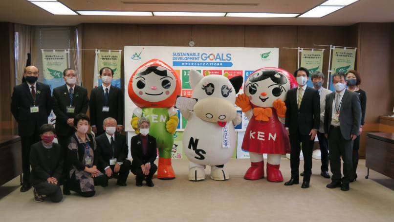 会議室の真ん中にみるひぃが立ち、左右に人KENまもるくんと人KENあゆみちゃんが立って、その周りをスーツを着た人々が囲むようにしている様子の集合写真