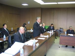 会議室で長机を前にして複数人が座っており、そのうちの1名が立ってマイクを手に話している写真