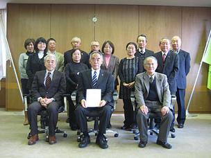 背後に2本の旗が置かれた会議室で、複数人の男女が正面を向いており、そのうちの真ん中の1名の男性が書物を手にしている集合写真