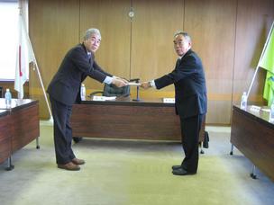 背後に2本の旗が置かれた会議室で、2人のスーツ姿の男性が向き合い、答申書の受け渡しをしている写真