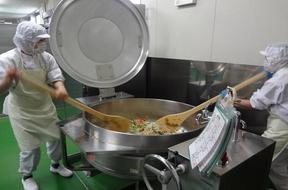 2人がかりで大きなヘラで窯の中の野菜を和えている様子の写真