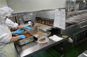 機械から出てくるフライ料理を受け取っている2人の白い作業着で身を包んだ職員の写真