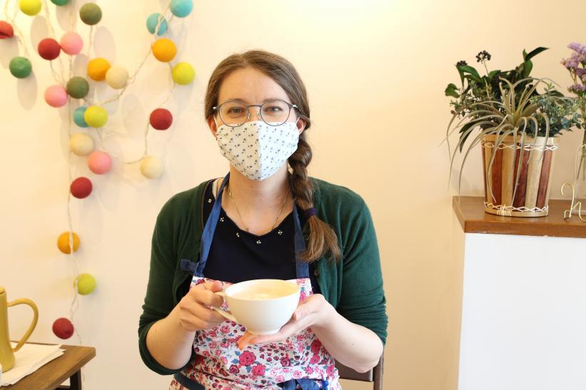 マスクとエプロン姿の女性が両手でコーヒーカップを持っている写真