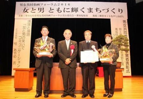 表彰式のステージで男性3人と女性一人が並んでいる写真