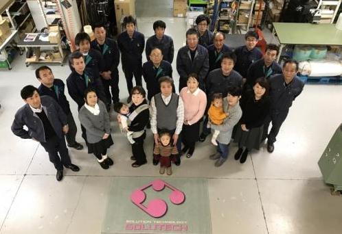 工場で男性が子供を抱えた女性何人かを囲むように立ち、正面斜め上を見上げている写真