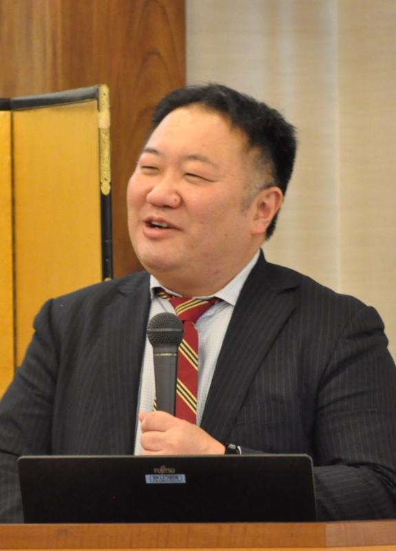 スーツを着た男性がマイクを持って話をしている写真