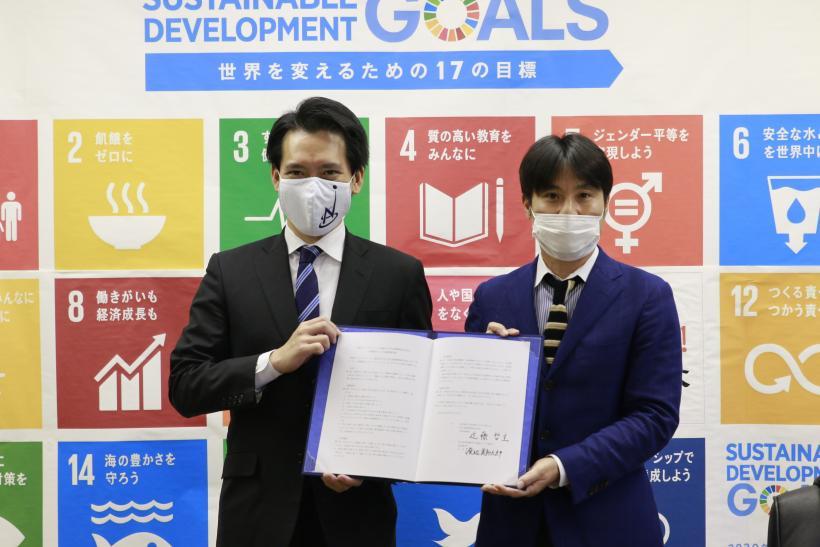 SDGsのポスターを背に、二人の男性が協定書を持ってこちらに見せている写真