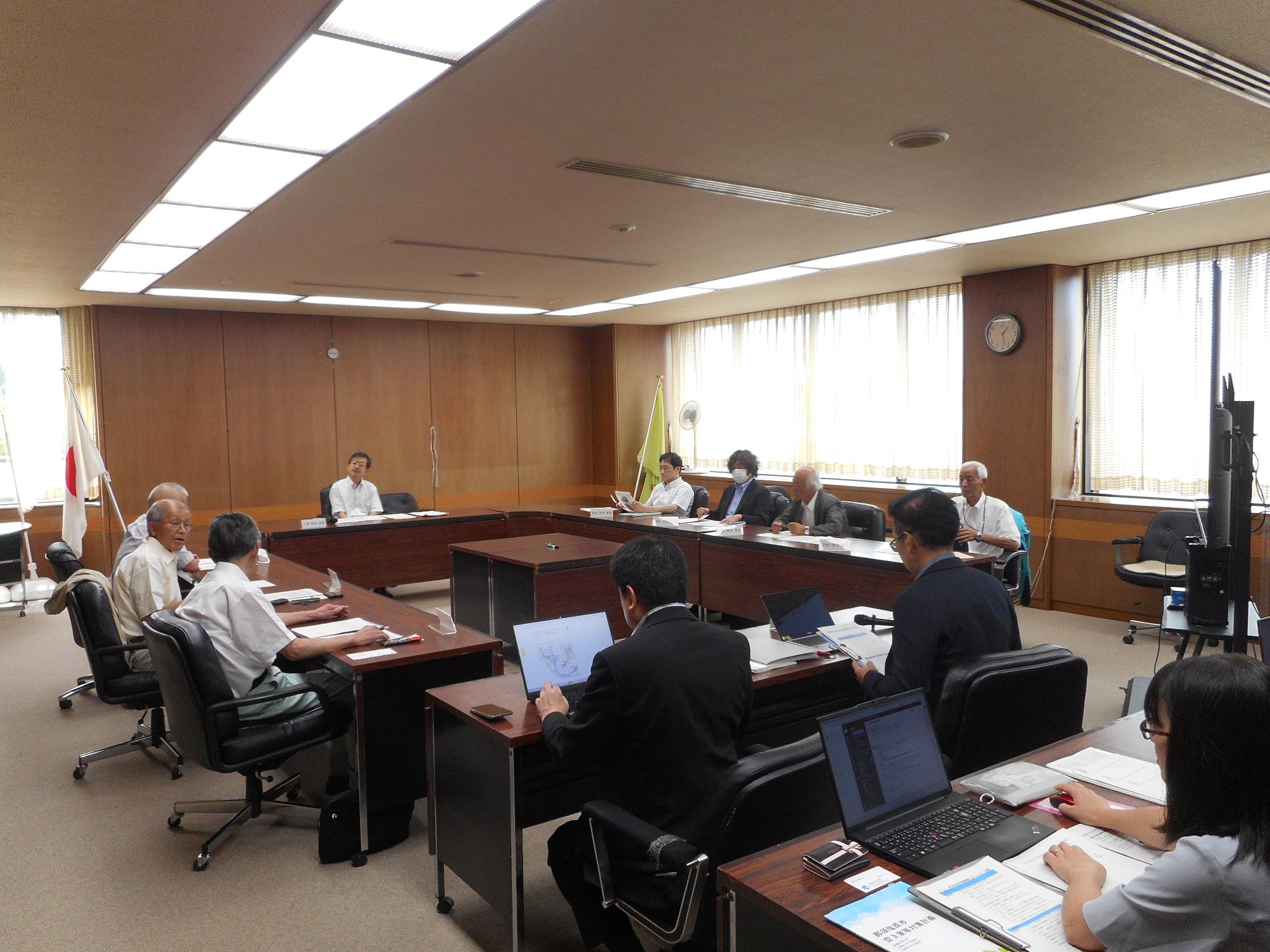 多くの人が長テーブルを四角に囲んで室内で審議会をしている写真
