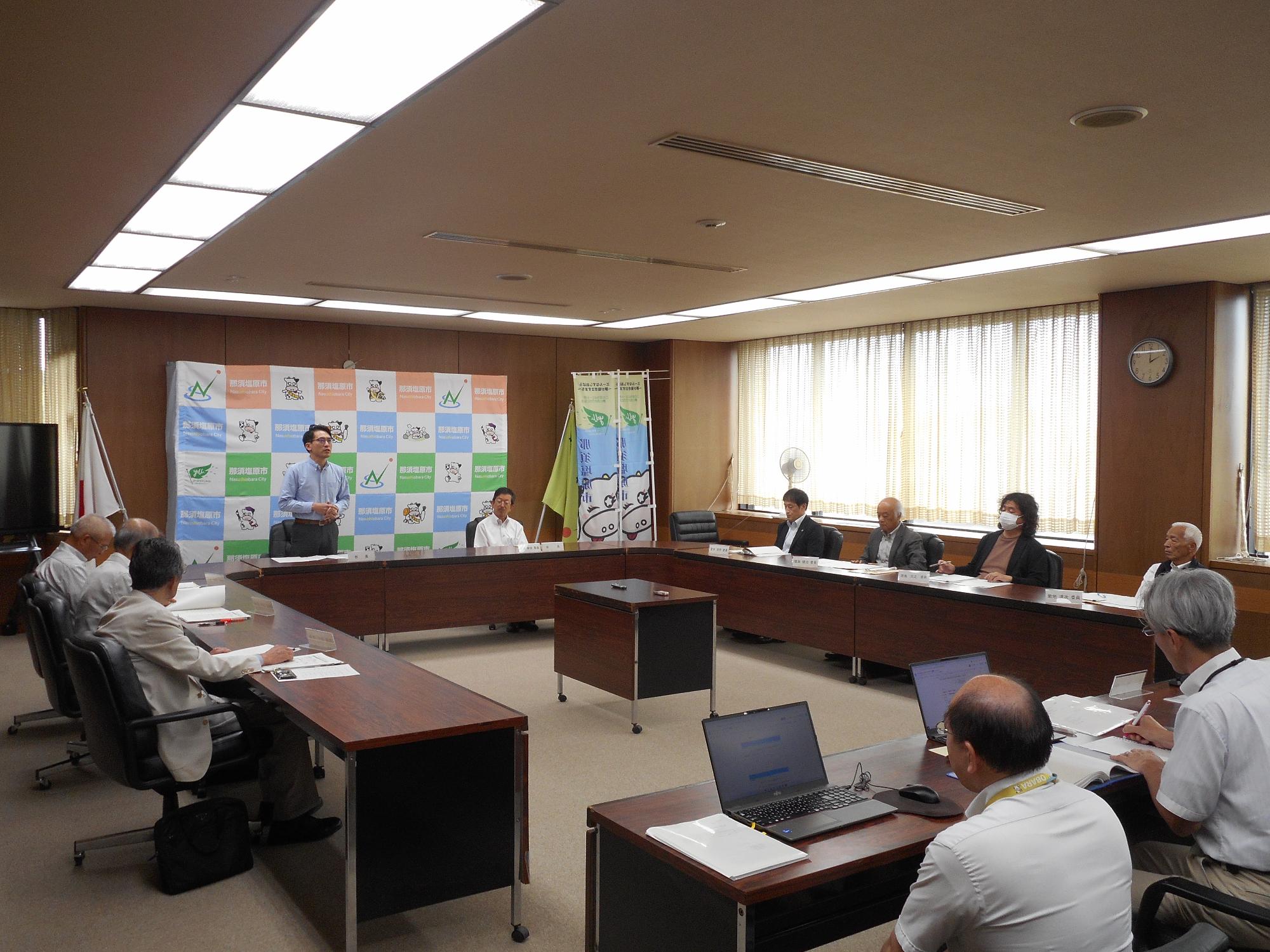 多くの人が長テーブルを四角に囲んで室内で審議会をしている写真