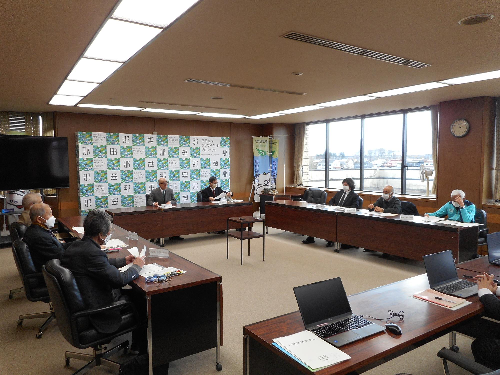 多くの人が長テーブルを四角に囲んで室内で審議会をしている写真