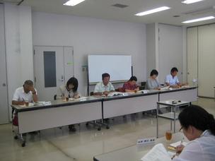 白い壁の明るい会議室で、6人の男女が長机を前にして横並びに座って書類を見ている写真