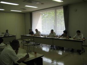 大きな窓から光が差し込む会議室で、複数の男女が長机を前にして座り書類を見ている写真