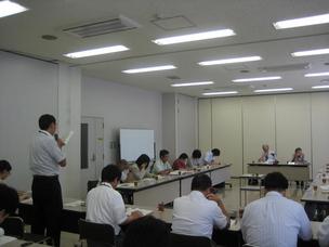 白い壁の明るい会議室で、複数の男女が長机を前にして座っており、そのうちの1名が書類を見ながらマイクを手にし話している写真