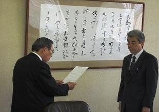 スーツ姿の男性がもう一人の男性に対して紙に書かれた文言を読み上げている写真