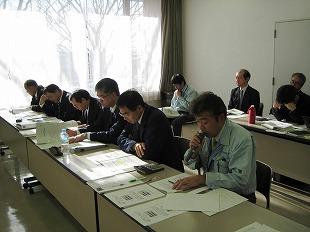 2列に並べられたテーブルの後ろに腰掛けている男性たちが手元の書類を見ている写真
