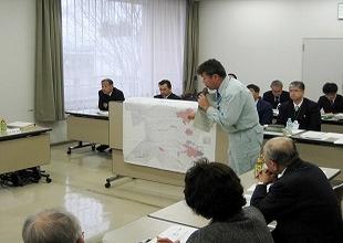 会議室でスーツ姿の複数の男女が長テーブルを囲んでおり、その中央で作業着を着た男性がマイクを手にし話している写真