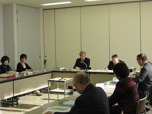 白い壁の会議室で、複数の男女が、目の前の長机に載せてある複数の書類を見ながら座って話し合っている写真