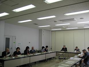 会議室で囲むように並べられた長机を前に複数の男女が座り手元の資料を眺めている写真