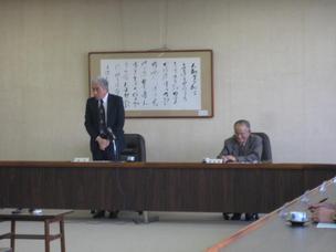 壁に額縁が貼られている会議室で、1人の男性が座って書類を見ていて、もう1人の男性が席を立って話をしている写真