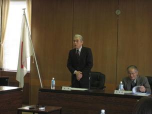 日本国旗が置かれた会議室で、一人のスーツ姿の男性が手元の資料を読んでおり、もう一人のスーツ姿の男性が立って話している写真