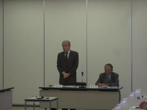 白い壁の会議室で1名の男性が座って書類を見ており、その隣にいる男性が立って挨拶をしている写真