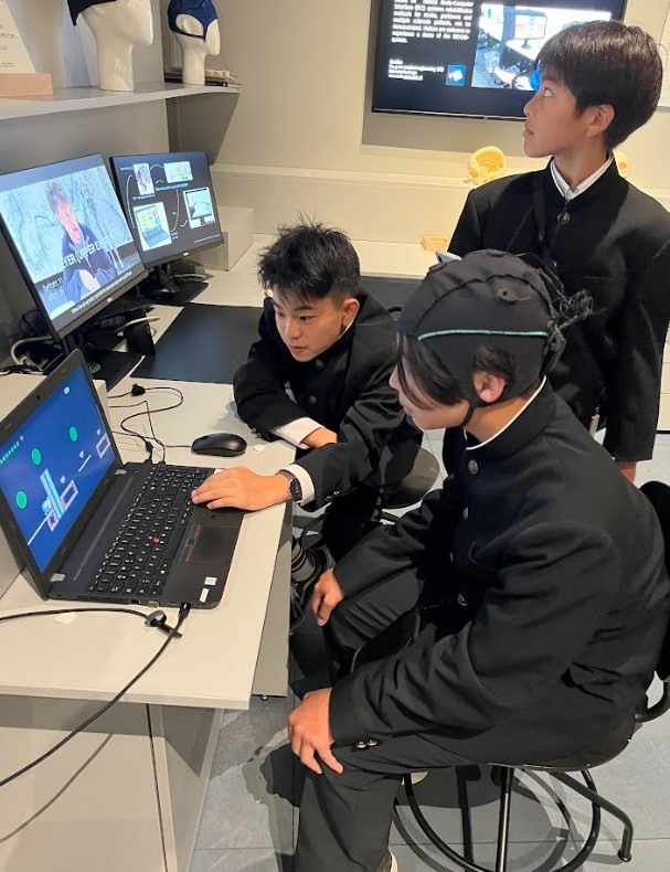 アルスエレクトロニカセンターで、学生服を着た3人の男子生徒が展示を体験している様子。1人の生徒が頭にセンサー付きのキャップを被り、脳波を使ってノートパソコンを操作しています。