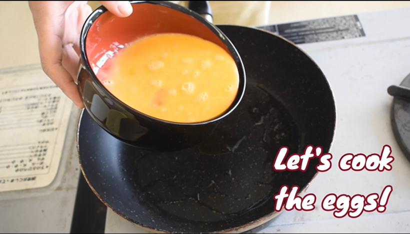 薄焼き卵を作る為、熱したフライパンに溶き卵を入れるシーンのサムネイル画像。画面右下には「卵を調理してみよう」と英語の字幕が表示されているのが確認できる