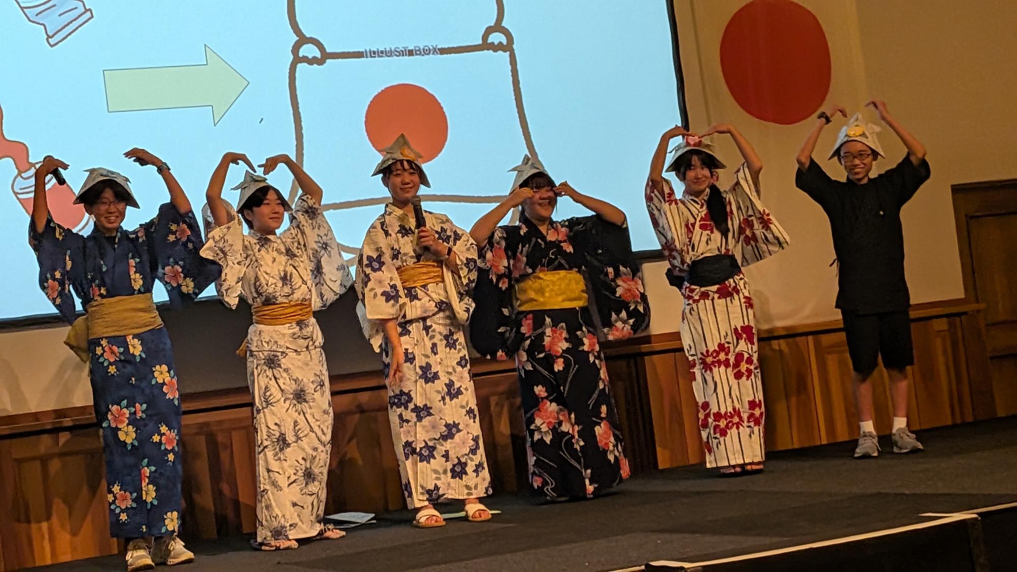 ステージ上で、浴衣姿などの生徒たちが頭に折り紙の兜をかぶり、両腕で頭の上にハートマークなどを作りながら、楽しそうに発表をしている様子。