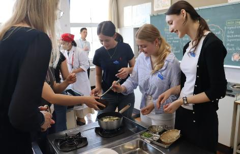家庭科室での調理実習の様子。数名のアウホフ校の生徒たちがコンロを囲み、笑顔で鍋からお椀へお玉で味噌汁を注いでいる。