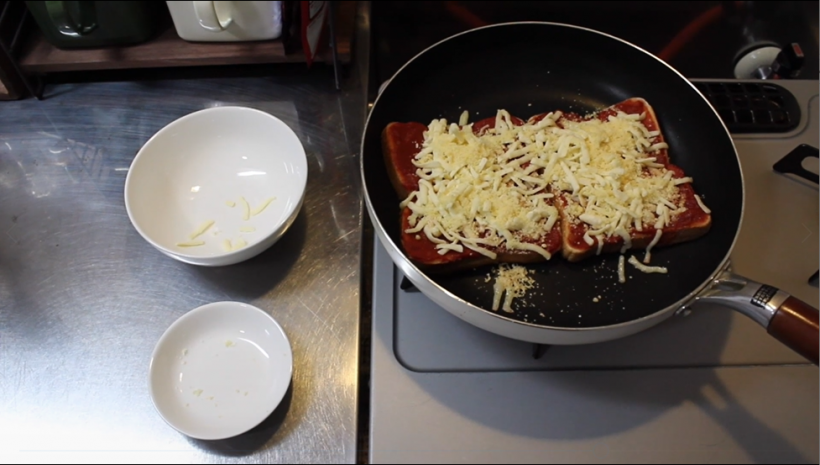 動画の中で、2枚のピザトーストをフライパンに並べてキッチンのガスコンロで焼いているシーンを切り取った写真。ガスコンロの横にはピザ用チーズと粉チーズが入っていた2枚の皿が置かれている。