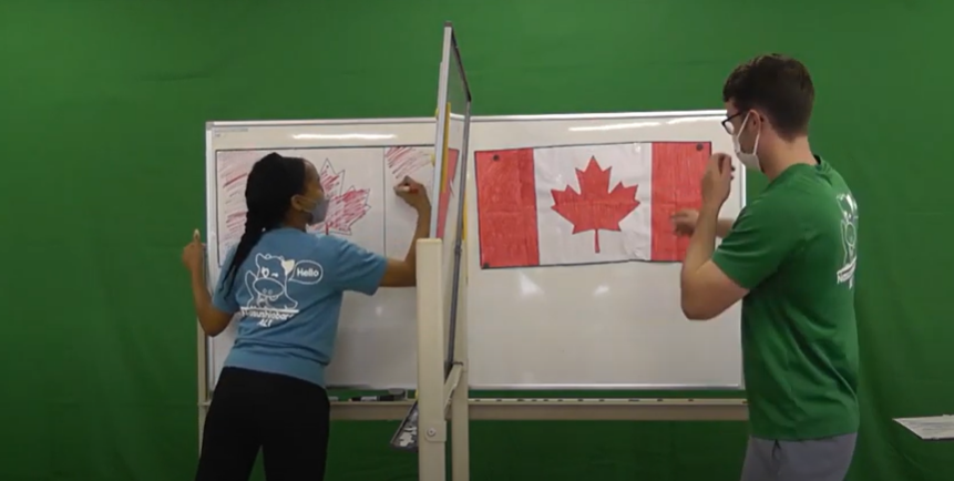 2名のALTがホワイトボードに向かっている。ホワイトボードは半分に分かれていて、カナダの国旗が二つあります。左側にいるALTは国旗を赤いマーカーペンを使って色を塗っている。国旗を完成させている。