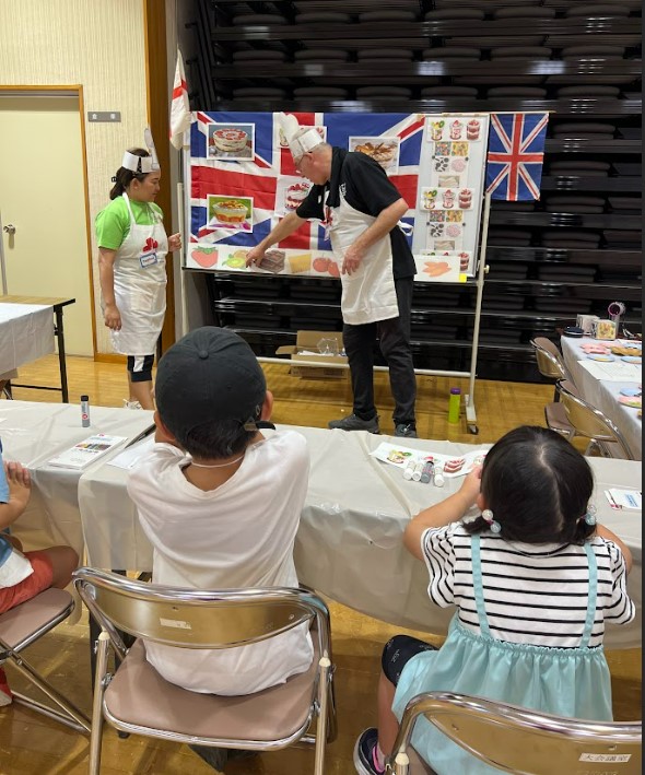 ALTが児童たちに果物の発音を教えている。ホワイトボードにはイギリスの国旗やくだもの絵カードが貼ってある。