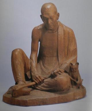 丸刈りの男性が片膝を立てて座りながら手作業をしている木造彫刻の写真