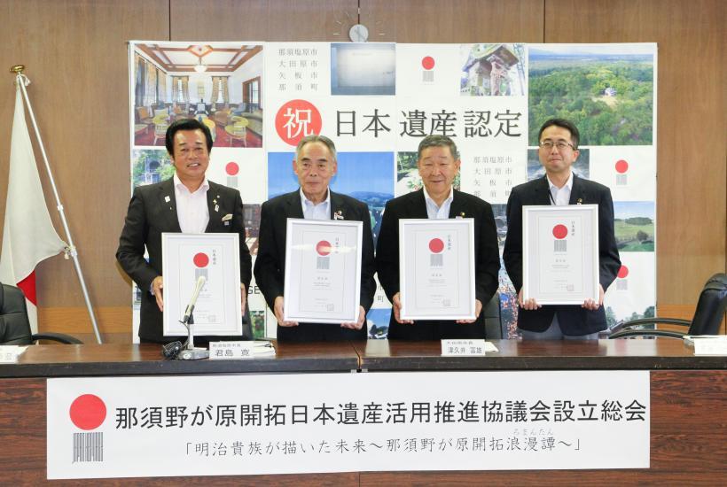 「那須野が原開拓日本遺産活用推進協議会設立総会」と書かれた紙が貼られた机の後ろに、額を持った男性4人が横に並んで立っている写真