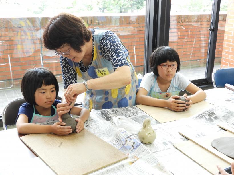 陶器の土鈴を作っている2人の女子小学生とそのお手伝いをしている先生の様子の写真