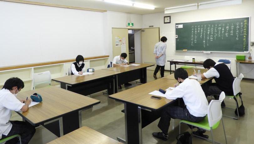 黒板前にコの字で机を並べそれぞれ紙に書き込んでいる5人の学生と、その周囲を歩く女性の写真