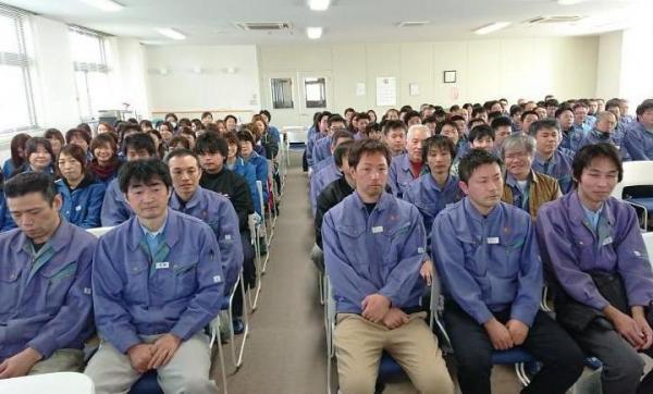 広い部屋に青い制服を羽織った人たちが沢山の列になって座る様子の金子メディックス株式会社の写真