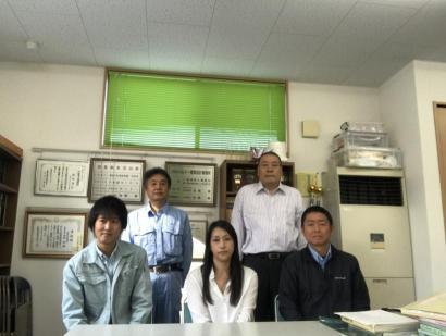 自社の表彰状を背景に並ぶ作業服やスーツ姿の男女の写真
