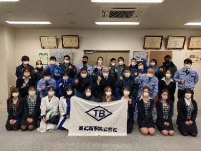表彰状が飾られた部屋で会社の旗を持ったりポーズをとったりする職員たちの写真