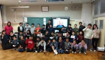 市立埼玉小学校の教室でおこなわれたオンライン交流会において、電子黒板に映し出されたシャッタウアー選手を囲んで記念撮影をする児童たちの写真