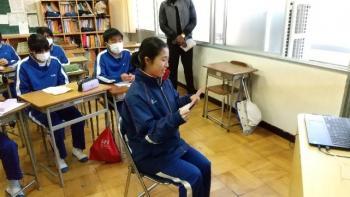 市立厚崎中学校の教室でおこなわれたオンライン交流会において、電子黒板の前の椅子に座ってメモを読みながら会話をする生徒の写真