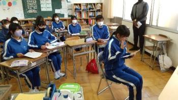 市立厚崎中学校の教室でおこなわれたオンライン交流会において、電子黒板の前の椅子に座って手の動きも加えながらメモを読みつつ会話をする生徒の写真