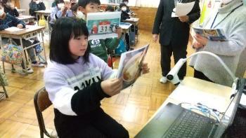 市立西小学校の教室でおこなわれたオンライン交流会において、通信機材である電子黒板の前で写真を使いながら日本の文化を紹介する児童たちの写真