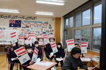 市立塩原小中学校の教室でおこなわれたオンライン交流会において、オーストリアの国旗と那須塩原市の市章が縦に並んだ棒を掲げる生徒たちの写真