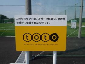 青木サッカー場Cグラウンド外縁に立つ、スポーツ振興くじの標識の写真。標識には「このグラウンドは、スポーツ振興くじ助成金を受けて整備されたものです」と書かれているのが確認できる
