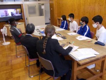 市立厚崎中学校の特別教室でおこなわれたオンライン交流会において、長机を囲んで座りながら電子黒板に映し出されたフリューヴィルト選手と会話をする生徒と学校関係者たちの写真