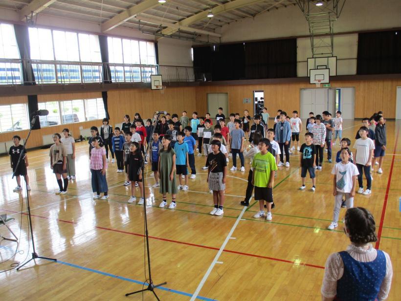 関谷小学校の体育館にて、距離を開けつつ身長よりも高い位置に設置された収録用マイクへ向けて合唱する児童たちの写真