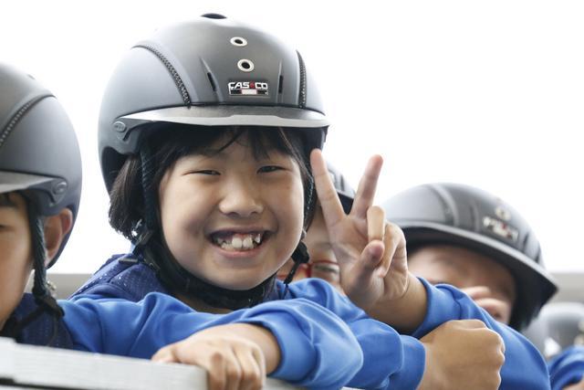 乗馬用ヘルメットを被って、満面の笑みとVサインをしつつ撮影に応じる児童の写真