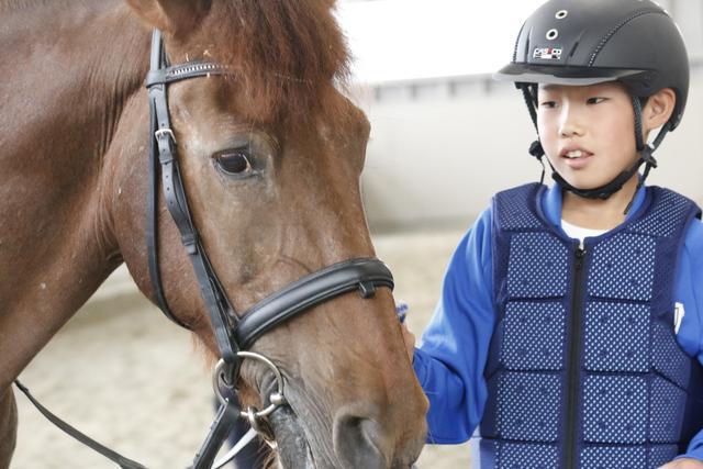 乗馬用ヘルメットとプロテクターを着用した姿で馬と触れ合う児童の写真