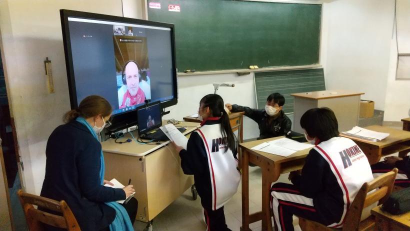 市立箒根中学校の教室でおこなわれたオンライン交流会にて、教室で机に電子黒板の前にあるパソコンのカメラの高さに合わせて立膝でバッハマイヤー選手と会話する生徒を左脇から撮影した写真
